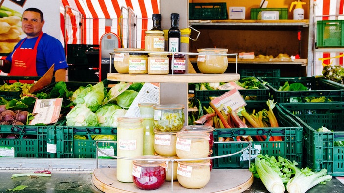Auf einem Aufsteller eines Marktstands stehen eingemachte Lebensmittel. Dahinter befinden sich ein Mitarbeiter des Standes und Kisten mit Gemüse.
