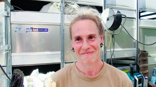 Eine Frau mit dunkelblonden Haaren und einem großen Ohrring lacht. Im Hintergrund ein Schafsgehege