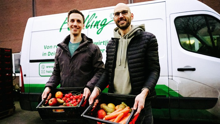 Johannes Becker links und Daniel Neumann rechts vor einem ihrer Gemüse-Transporter. Beide halten eine Gemüsekiste in der Hand.