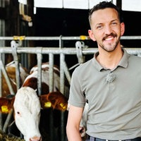 Ein Mann steht mit einen Polohemd bekleidet in einem Kuhstall, hinter ihm sind Kühe zu sehen, die gerade fressen