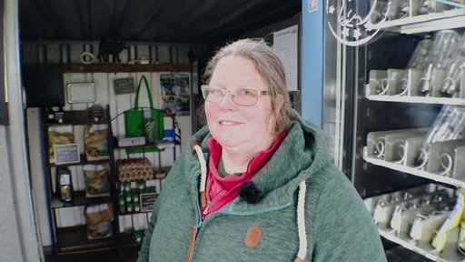 Eine Frau mit Brille schaut in die Kamera. Sie trägt eine grüne Jacke. Im Hintergrund ist ein Hofladen.