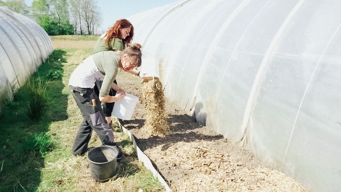 Zwei Frauen stehen vor einem Beet und streuen Mulch darüber.