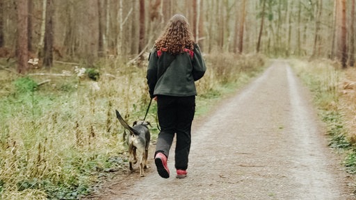 Eine Frau mit lockigen, braunen Haaren von hinten. Sie läuft mit einem Hund einen schmalen Weg in einem Wald entlang.