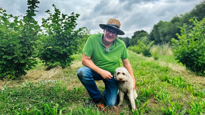 Burkhard Kleffmann, grünes Polo-Shirt, Strohhut, Brille, blaue Jeans, hockt im Gras rechts neben ihm sitzt seine Hündin Betty.