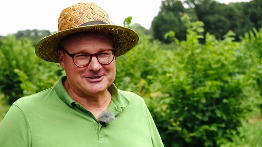 Burkhard Kleffmann, Strohhut, rote Brille, grüßes Polo-Shirt, im Hintergrund grüne Bäume und Pflanzen