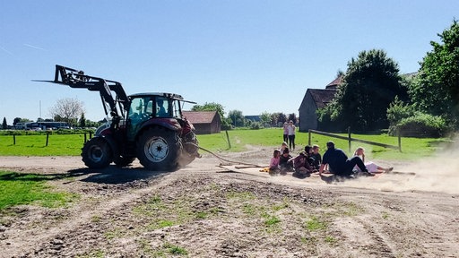 Ein grüner Trecker verbunden mit einem, auf dem Boden liegenden Teppich. Darauf einige Kinder und eine erwachsene Person. Im Hintergrund eine grüne Weide