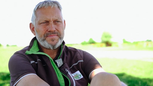 Ein Mann mit kurzen grauen Haaren, Bart und einer grau-grünen Fließjacke. Im Hintergrund eine grüne Weide
