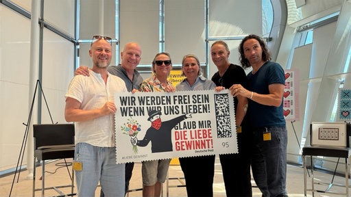 Die Band Brings, fünf Männer mittleren Alters, halten eine große Briefmarke zusammen mit einer Frau.