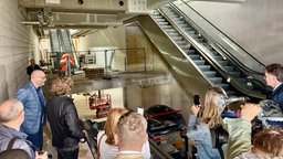 Das ehemalige Kaufhofgebäude von innen. Rechts sieht man zwei Rolltreppen, im Hintergrund die unverputzten Wände. Vorne stehen die Journalisten.