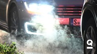 Autos stehen im Stau und stoßen Abgase in die Luft. Es ist dunkel und die Scheinwerfen des hinteren Autos leuchten hell. 