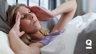 Eine Frau mit blonden Haaren liegt im Bett und hält ihre Hände an ihren Kopf. 