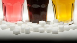 Drei Gläser gefüllt mit roter Limonade, Cola und einem Energy-Drink stehen neben Zuckerwürfeln auf einem Tisch.