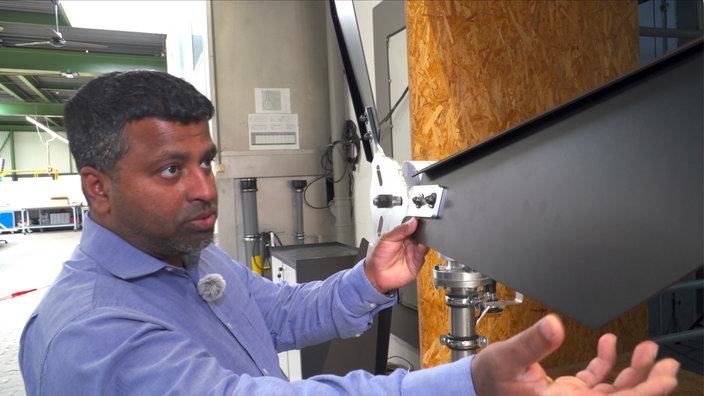 Ingenieur Sawkat Hossain steht in einem blauen Hemd  in einer Fabrikhalle, in der Hand hält er ein dunkles Bauteil