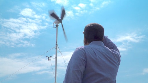 Ein Mann in einem blauen Hemd steht vor einem Windrad, das sich im Wind dreht und blickt mit erhobenem Kopf in Richtung Propeller