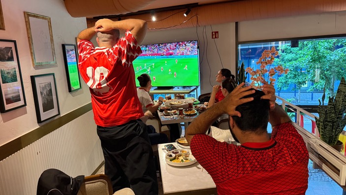 Fußballfans schauen gemeinsam das WM-Spiel. Die hinteren beiden Personen stehen mit dem Rücken zur Kamera und halten sich die Hände an den Kopf.