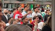 Drei junge Männer stehen zwischen weiteren Fußballfans und schauen das WM-Spiel 
