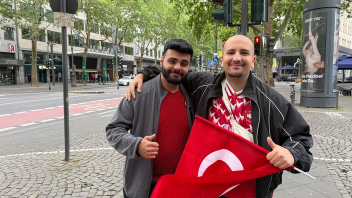 Zwei nebeneinanderstehende Männer lächeln in die Kamera. Die rechte Person hält eine Türkei-Flagge in der Hand.